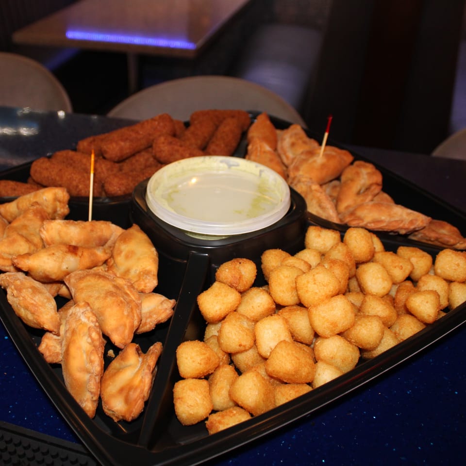 A black platter of Cuban appetizers including croquettes, empanadas, and fried cheese balls with dip.