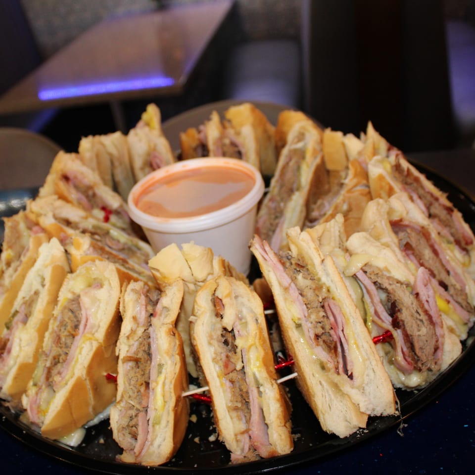 A platter of sliced Cuban sandwiches with ham, pork, cheese, and pickles, served with a dipping sauce.